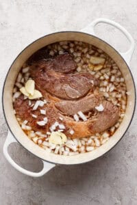 A large piece of beef is cooking in a white Dutch oven with chopped onions, garlic cloves, and liquid on a light textured surface.