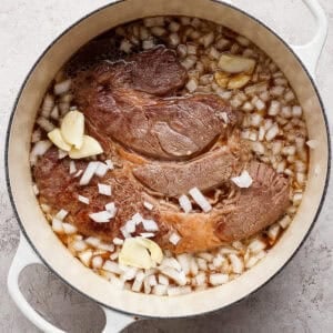 A large piece of beef is cooking in a white Dutch oven with chopped onions, garlic cloves, and liquid on a light textured surface.