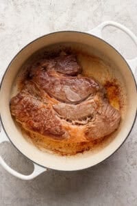 A large piece of seared beef sits in a white Dutch oven on a light-colored surface.