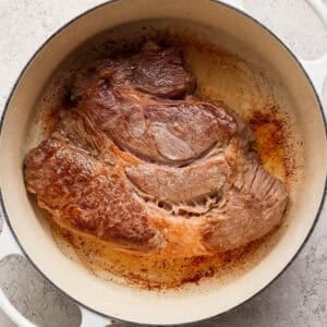 A large piece of seared beef sits in a white Dutch oven on a light-colored surface.