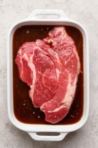 A raw beef steak sits in a white ceramic dish filled with brown marinade on a light textured surface.
