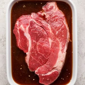 A raw beef steak sits in a white ceramic dish filled with brown marinade on a light textured surface.