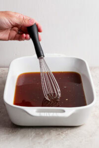 A hand uses a whisk to mix a dark liquid marinade in a white rectangular baking dish on a light surface.