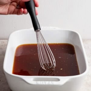 A hand uses a whisk to mix a dark liquid marinade in a white rectangular baking dish on a light surface.