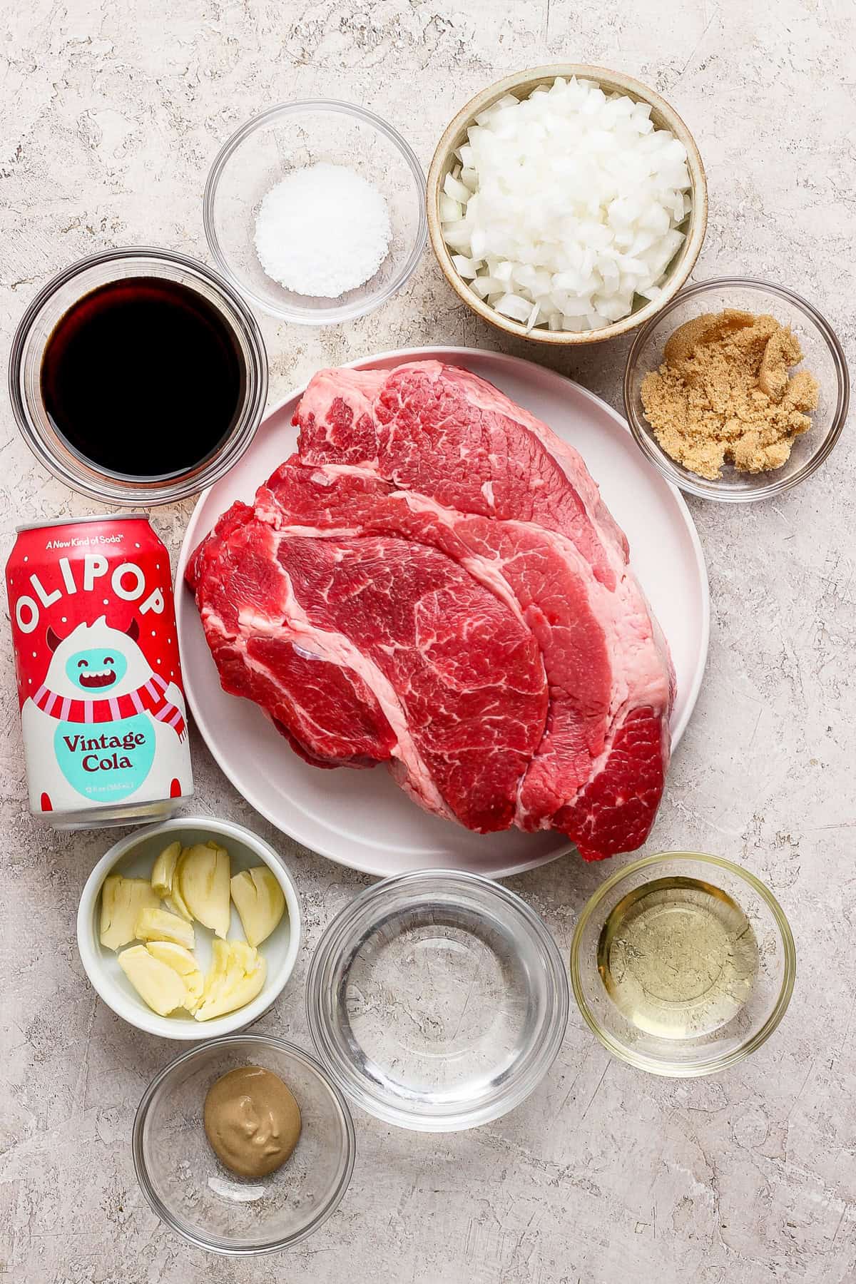Raw chuck roast on a plate, surrounded by bowls of chopped onions, brown sugar, garlic, salt, vinegar, mustard, oil, soy sauce, and a can of OLIPOP Vintage Cola on a light surface.