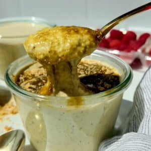 A spoon lifts thick, creamy pudding from a glass jar, with crumbs and a dark sauce on top; fresh raspberries are visible in the background.