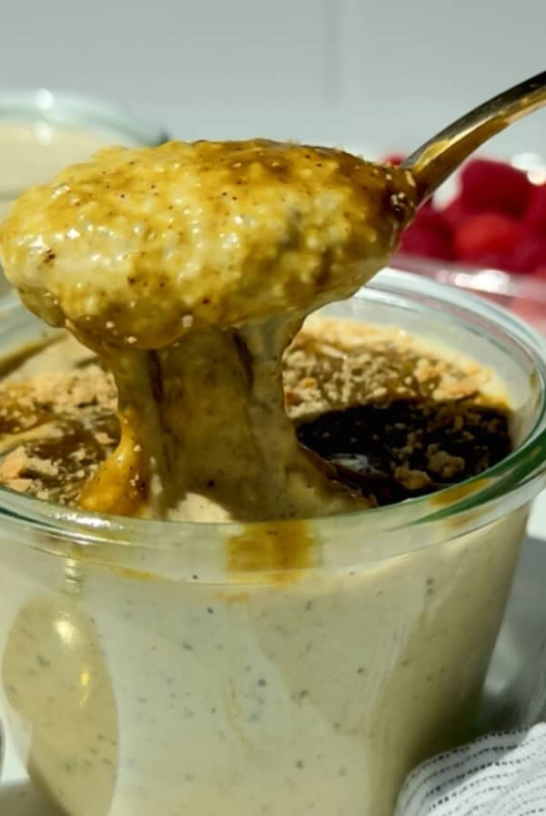 A spoon lifts thick, creamy pudding from a glass jar, with crumbs and a dark sauce on top; fresh raspberries are visible in the background.