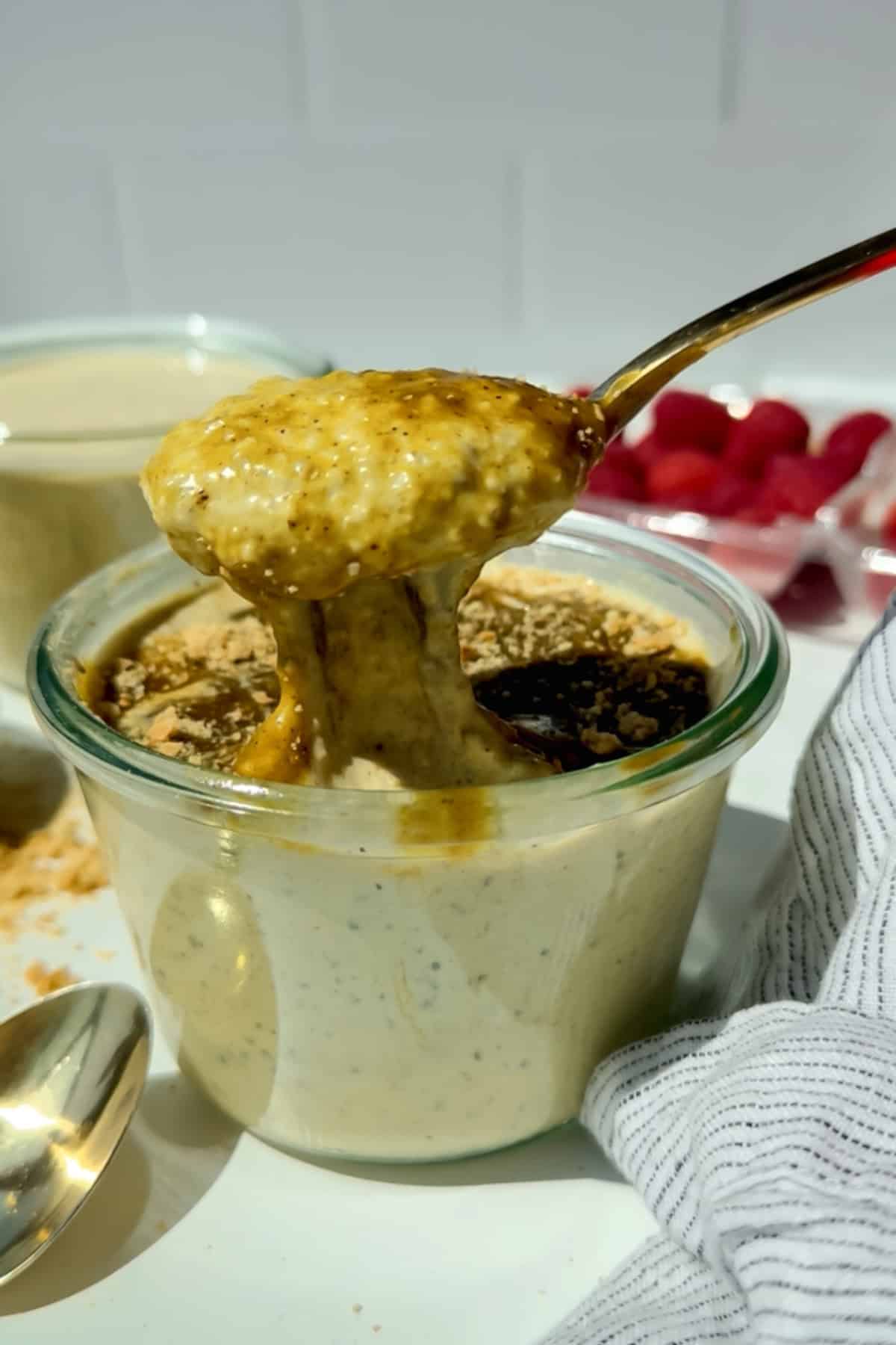 A spoon lifts creamy overnight oats from a glass jar, topped with a drizzle of syrup and a sprinkle of crumbs, with raspberries and a striped cloth nearby.