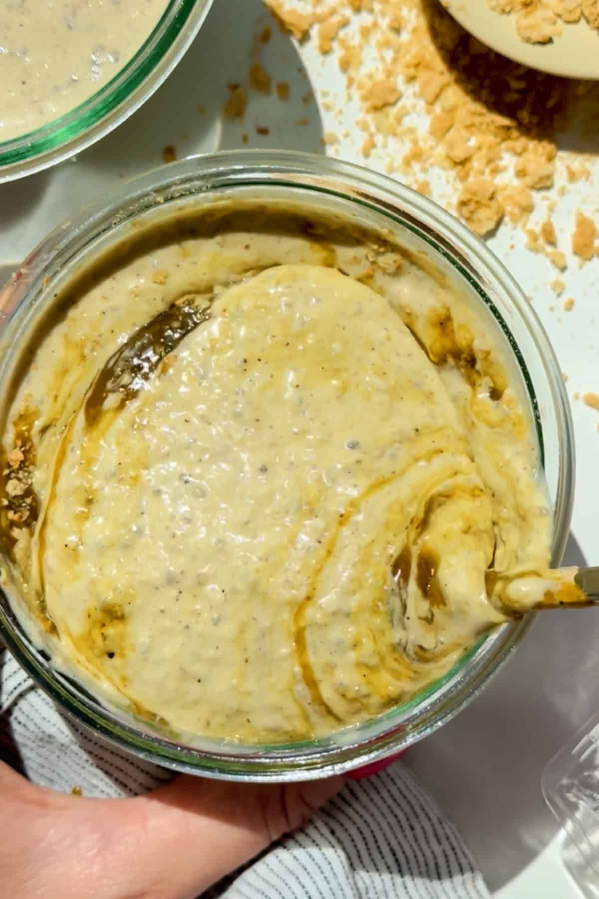 A glass bowl containing a creamy, beige mixture with swirls and specks, being stirred with a spoon. Crumbled pieces are scattered nearby on a white surface.
