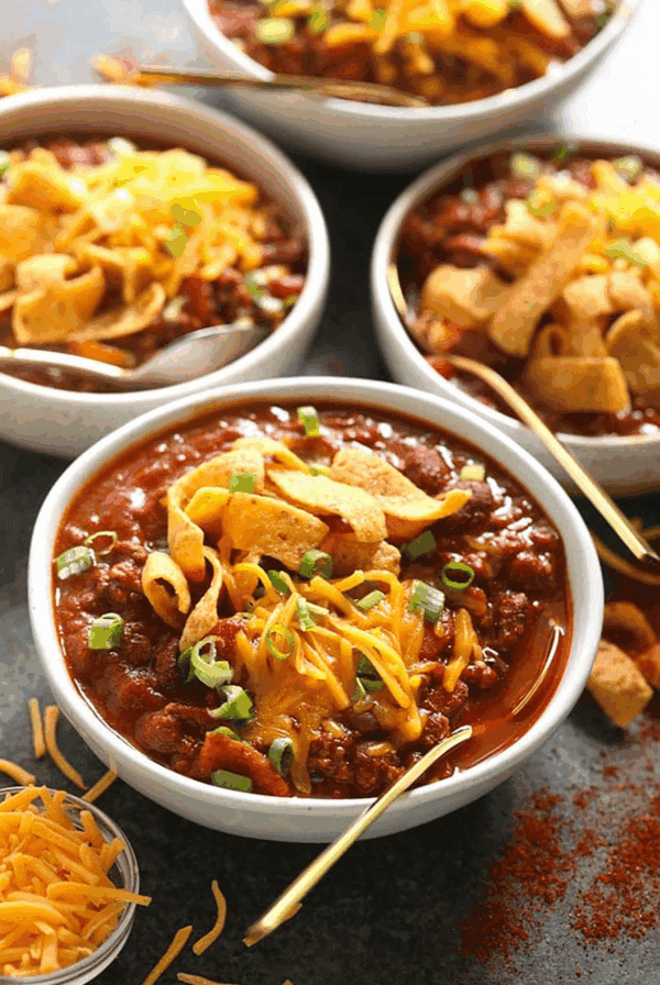 Four bowls of chili topped with shredded cheddar cheese, corn chips, and chopped green onions, with a side of shredded cheese and gold spoons.