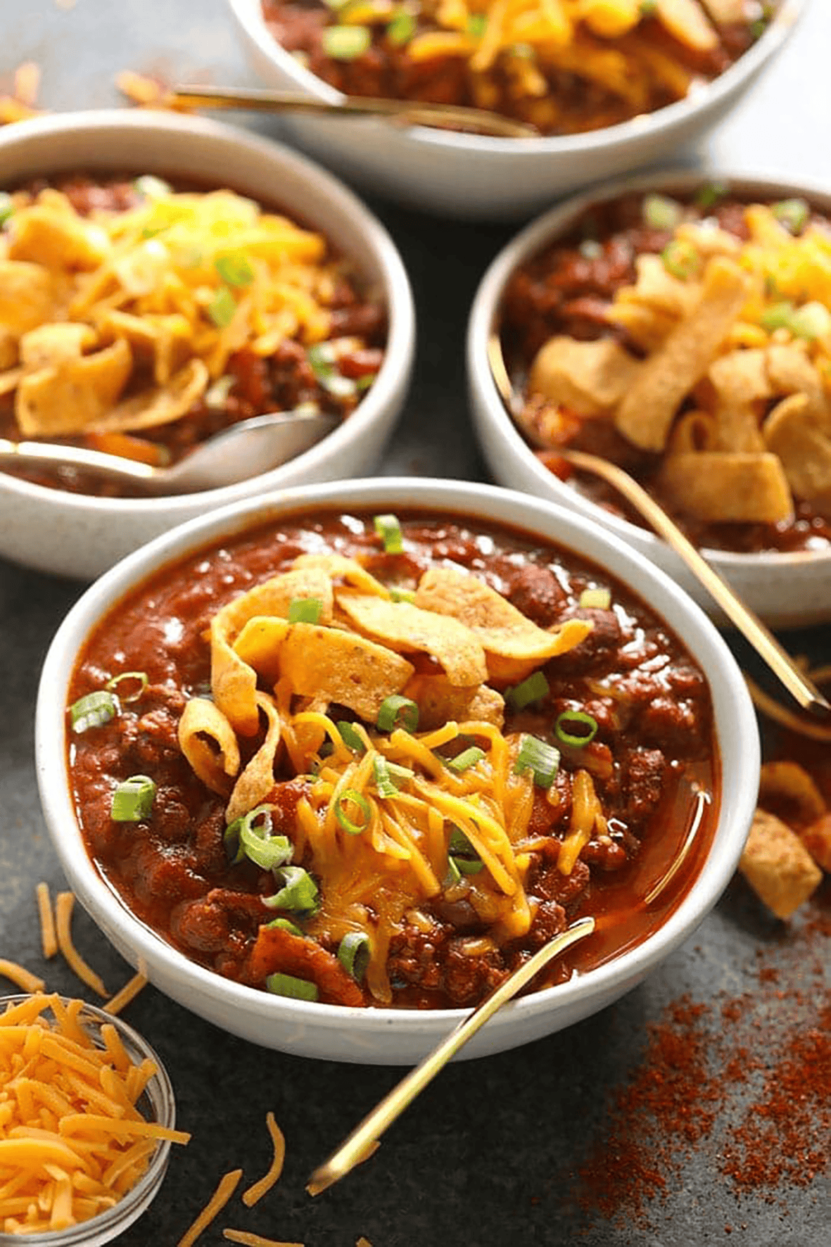 Four bowls of chili topped with shredded cheddar cheese, corn chips, and chopped green onions, with a side of shredded cheese and gold spoons.