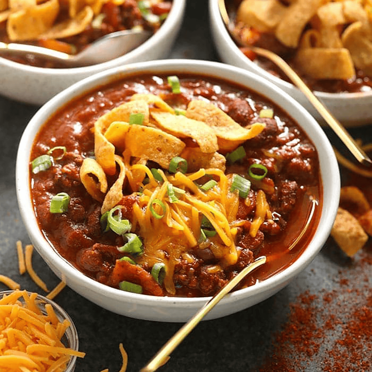 A bowl of chili recipe topped with shredded cheddar cheese, corn chips, and chopped green onions, served with a gold spoon and extra bowls in the background.