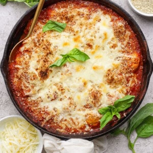 A cast iron skillet filled with baked cheesy tomato dish, garnished with fresh basil, surrounded by small bowls of shredded cheese and breadcrumbs.