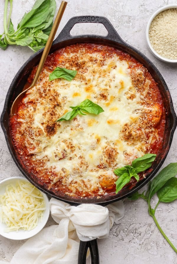 A cast iron skillet filled with baked cheesy tomato dish, garnished with fresh basil, surrounded by small bowls of shredded cheese and breadcrumbs.