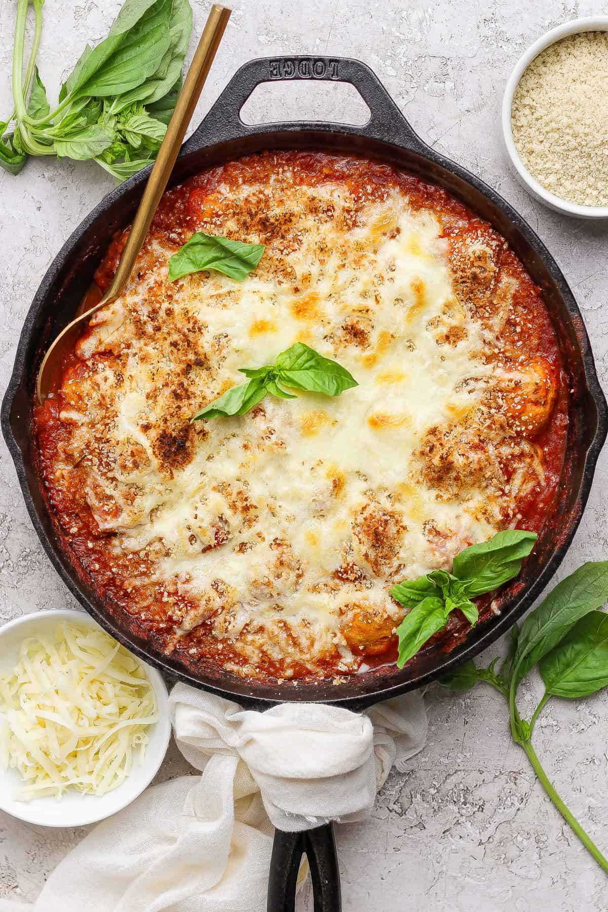 A cast iron skillet filled with baked cheesy tomato dish, garnished with fresh basil, surrounded by small bowls of shredded cheese and breadcrumbs.