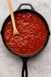A cast iron skillet filled with chunky tomato sauce, with a wooden spoon resting inside, on a light textured surface.