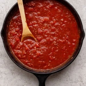 A cast iron skillet filled with chunky tomato sauce, with a wooden spoon resting inside, on a light textured surface.