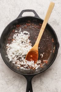 Chopped onions being sautéed in a cast iron skillet with a wooden spoon; residue from previous cooking visible in pan.