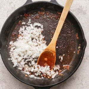 Chopped onions being sautéed in a cast iron skillet with a wooden spoon; residue from previous cooking visible in pan.
