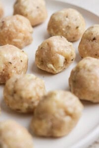 Raw chicken meatballs with visible herbs and cheese pieces arranged on a white plate, ready to be cooked.