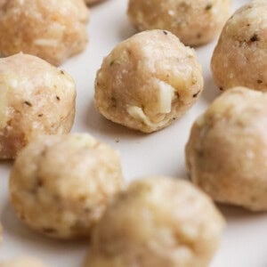 Raw chicken meatballs with visible herbs and cheese pieces arranged on a white plate, ready to be cooked.