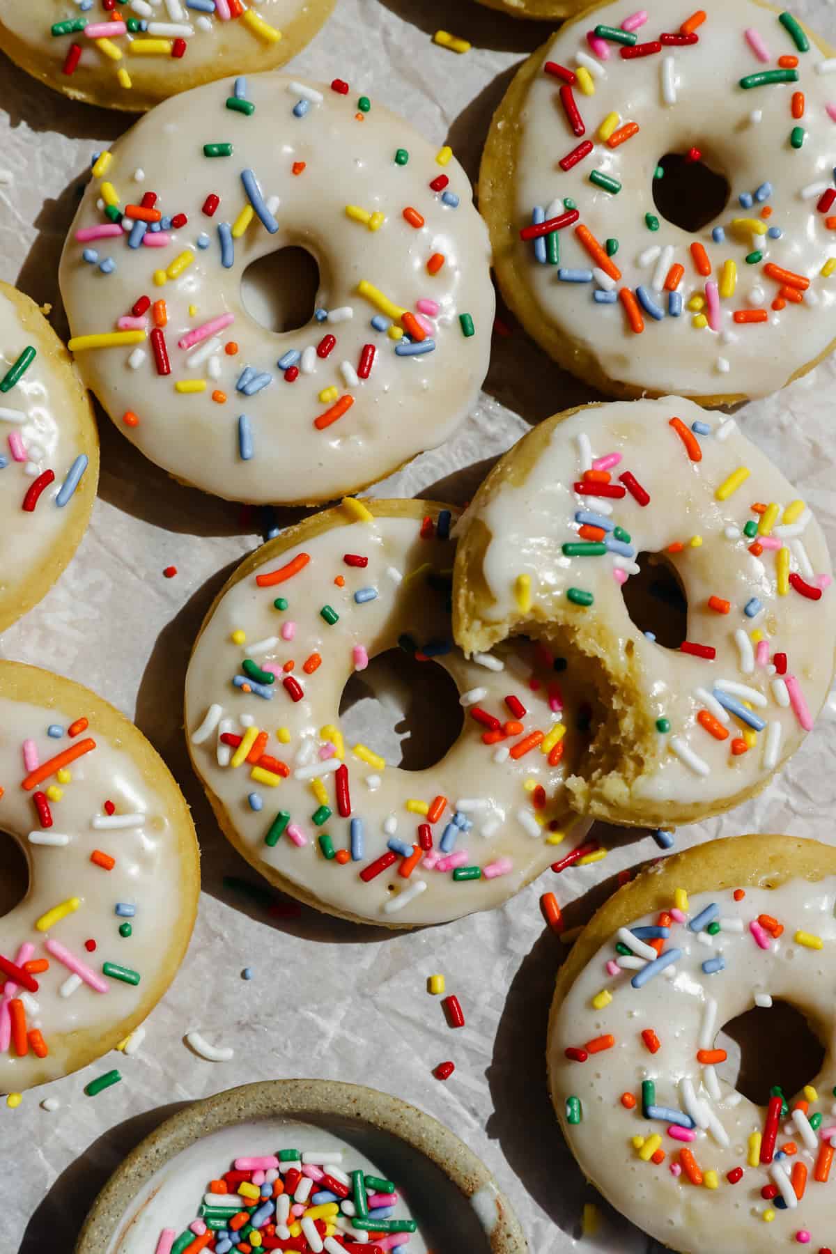 Glazed donuts topped with colorful sprinkles are arranged on parchment paper, with one donut partially eaten and a small bowl of extra sprinkles nearby.