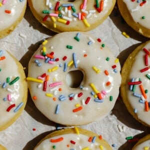 A close-up of vanilla-frosted donuts topped with colorful sprinkles arranged on parchment paper.