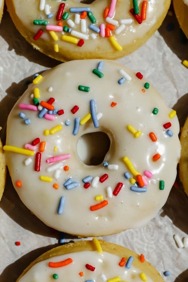 A close-up of vanilla-frosted donuts topped with colorful sprinkles arranged on parchment paper.