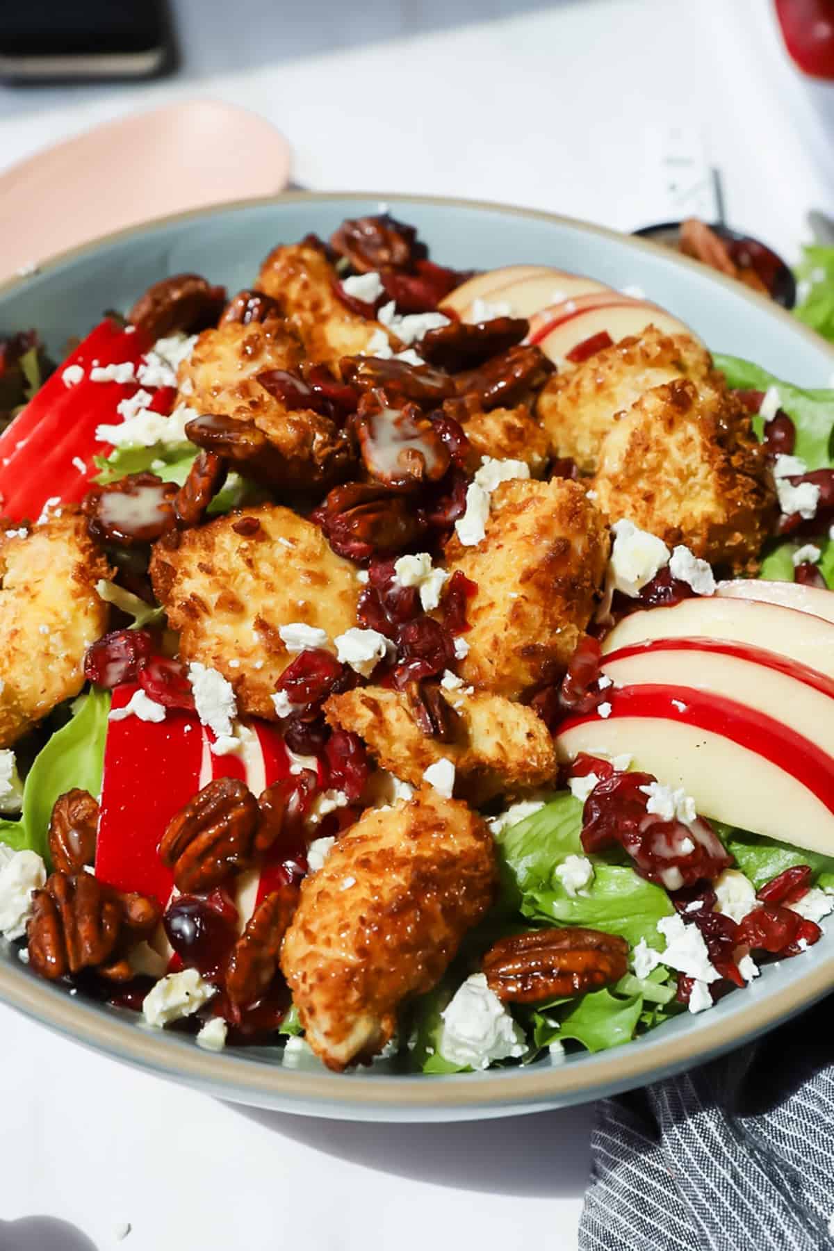 A salad with fried chicken pieces, sliced apples, pecans, dried cranberries, crumbled cheese, and mixed greens in a bowl.