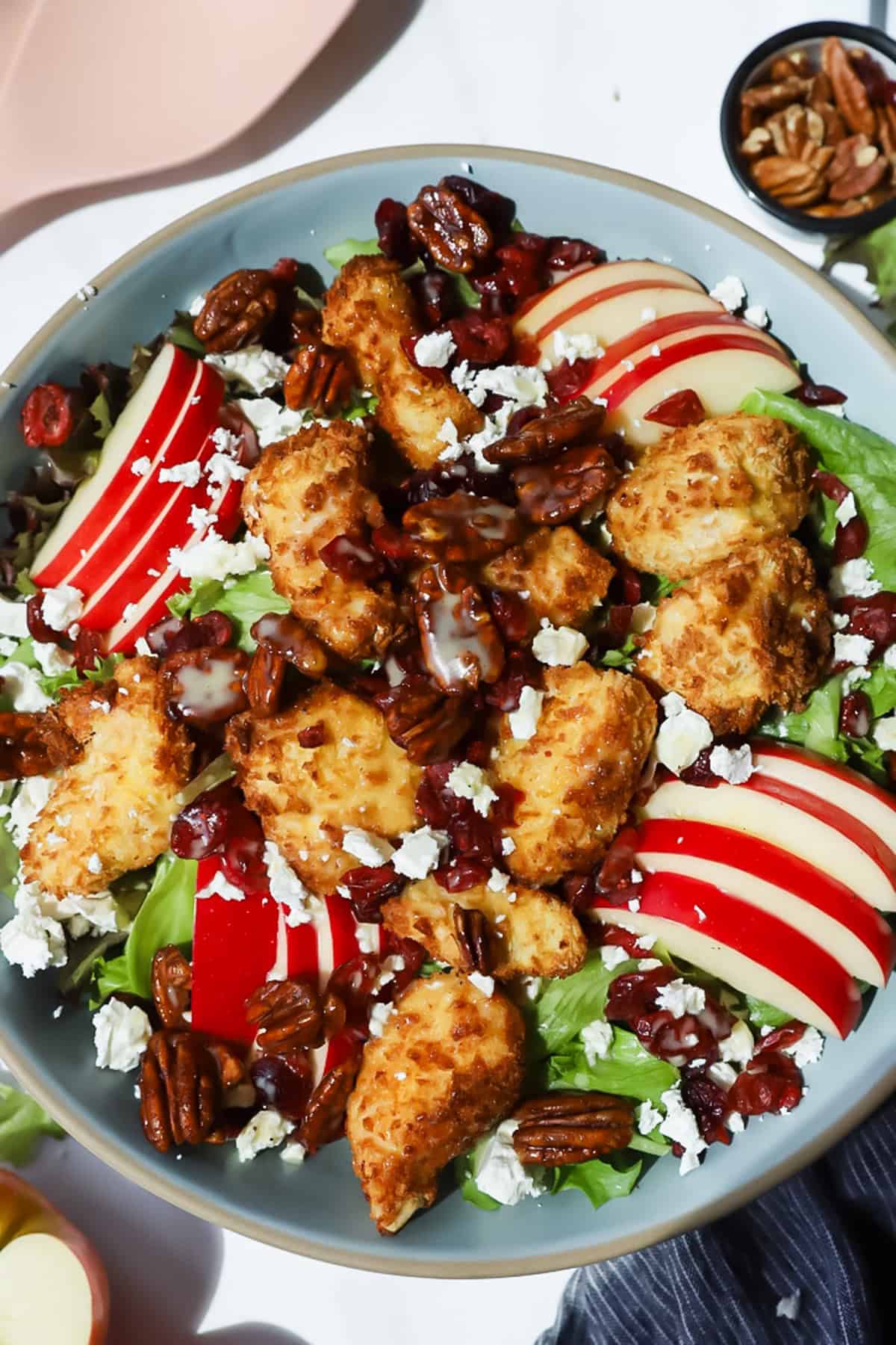 A salad with breaded chicken pieces, sliced red apples, pecans, dried cranberries, lettuce, and crumbled cheese, drizzled with dressing in a gray bowl.