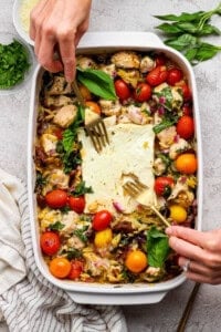 A person uses two forks to serve a baked dish with chicken, cherry tomatoes, greens, orzo, and feta cheese in a white casserole dish. Fresh herbs are visible on the side.