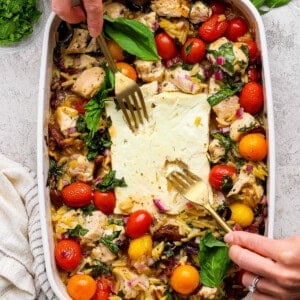 A person uses two forks to serve a baked dish with chicken, cherry tomatoes, greens, orzo, and feta cheese in a white casserole dish. Fresh herbs are visible on the side.