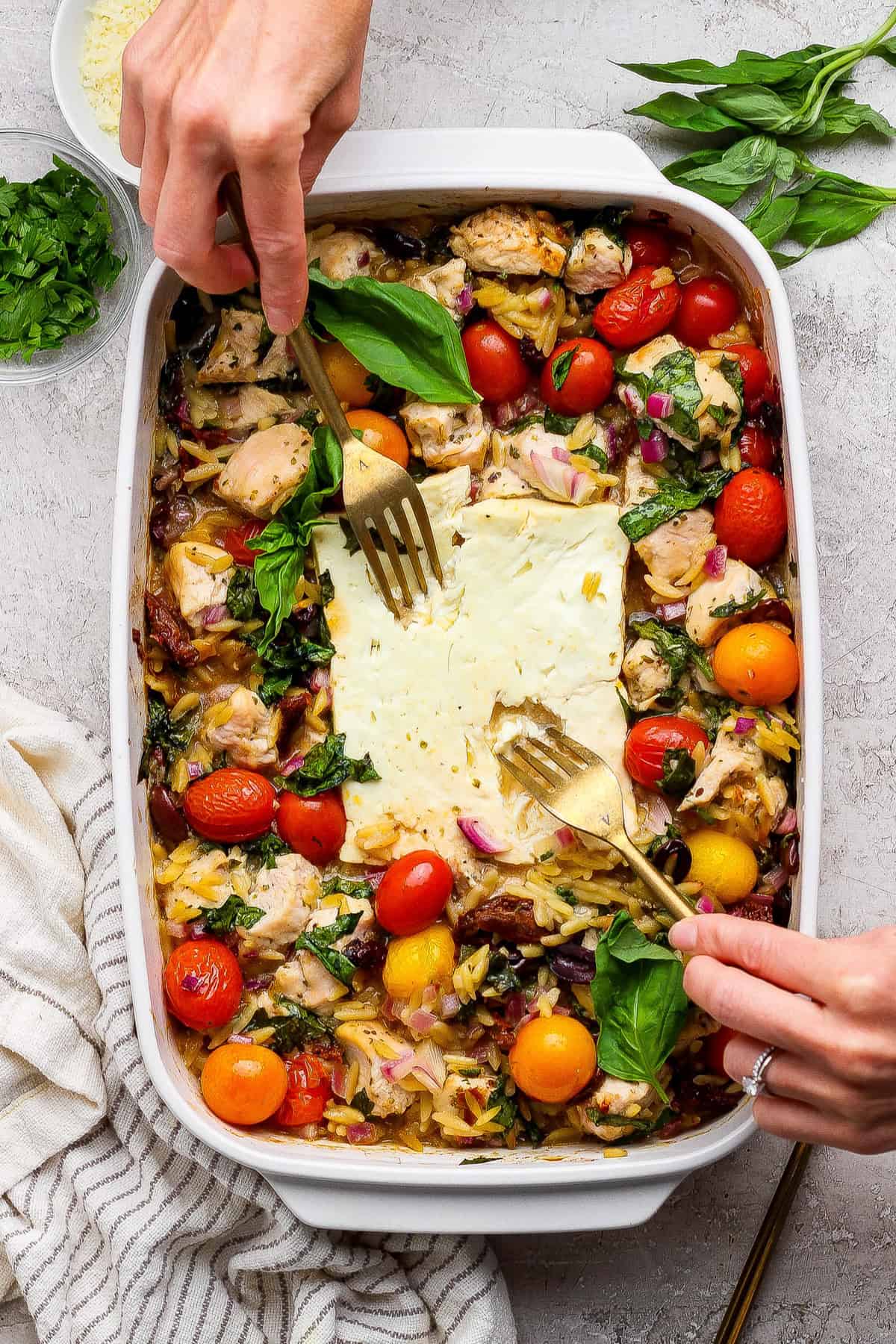 A person uses two forks to serve a baked dish with chicken, cherry tomatoes, greens, orzo, and feta cheese in a white casserole dish. Fresh herbs are visible on the side.
