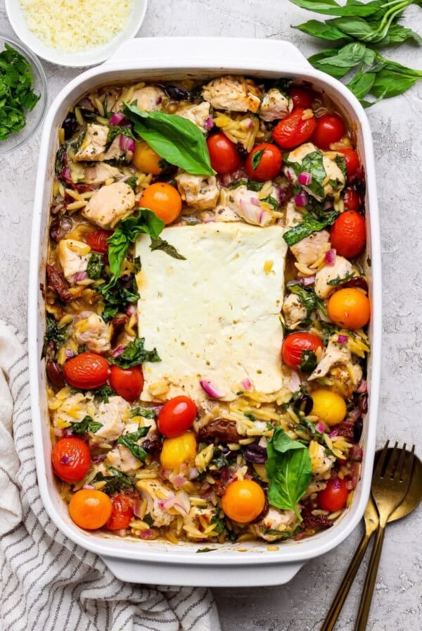 A baked casserole dish filled with chicken, cherry tomatoes, orzo pasta, red onions, and a large block of feta cheese in the center, garnished with fresh basil.