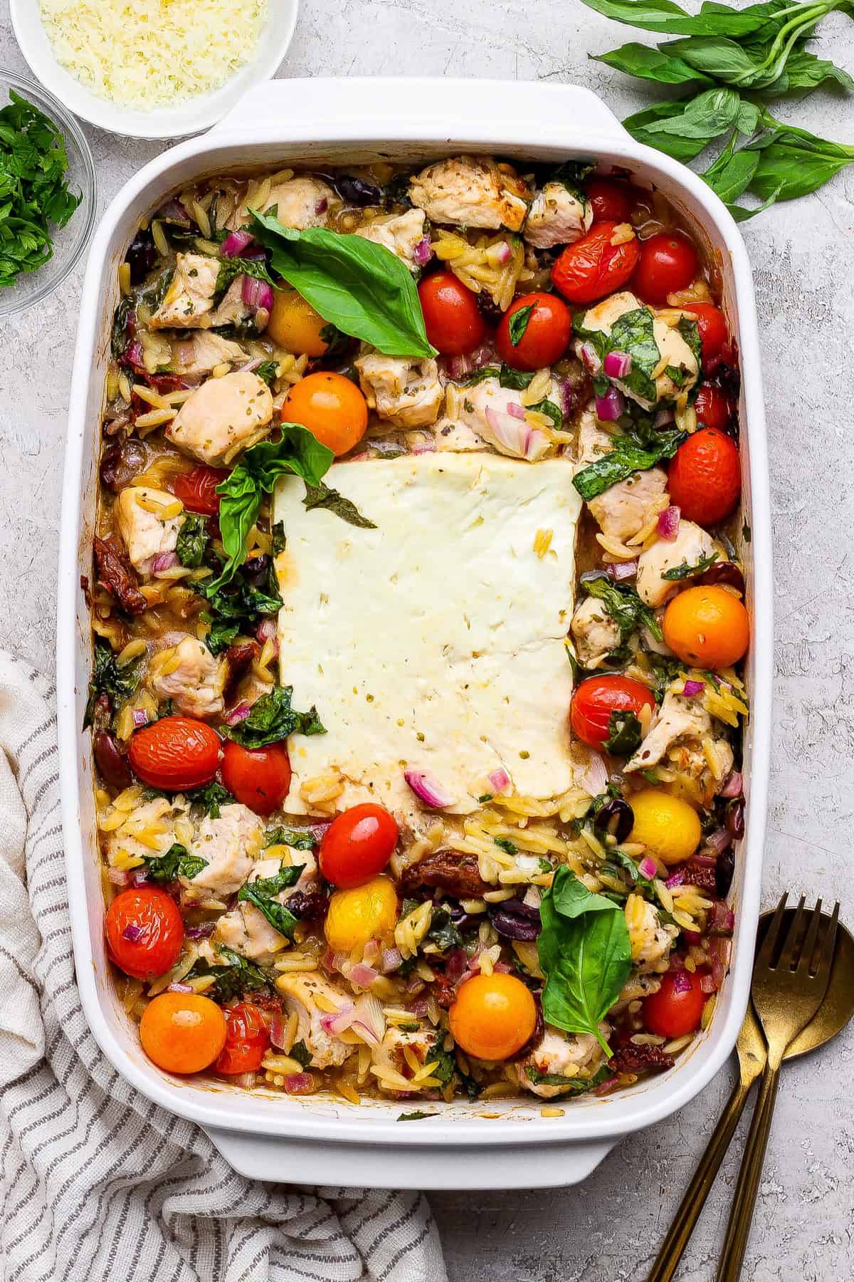 A baked casserole dish filled with chicken, cherry tomatoes, orzo pasta, red onions, and a large block of feta cheese in the center, garnished with fresh basil.