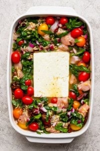 A white baking dish filled with raw chicken pieces, cherry tomatoes, spinach, chopped red onions, orzo, and a large block of feta cheese in the center.