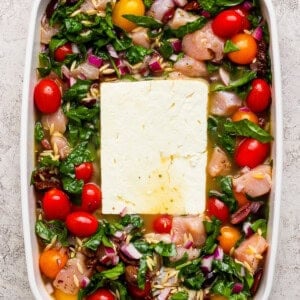 A white baking dish filled with raw chicken pieces, cherry tomatoes, spinach, chopped red onions, orzo, and a large block of feta cheese in the center.