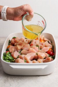 A hand pours broth from a glass measuring cup over raw chopped chicken, tomatoes, and greens in a white baking dish.