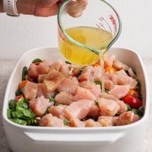 A hand pours broth from a glass measuring cup over raw chopped chicken, tomatoes, and greens in a white baking dish.
