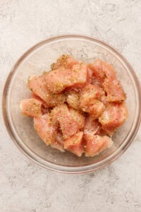 A glass bowl filled with raw, diced chicken breast pieces, seasoned with salt, pepper, and dried herbs, on a light textured surface.