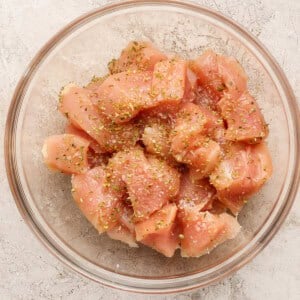 A glass bowl filled with raw, diced chicken breast pieces, seasoned with salt, pepper, and dried herbs, on a light textured surface.