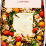 A baked Greek chicken orzo casserole topped with cherry tomatoes, spinach, herbs, and a large slice of feta cheese in a white dish.