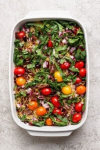 A white baking dish filled with fresh spinach, cherry tomatoes, chopped red onion, sun-dried tomatoes, and olives on a light textured surface.