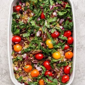 A white baking dish filled with fresh spinach, cherry tomatoes, chopped red onion, sun-dried tomatoes, and olives on a light textured surface.