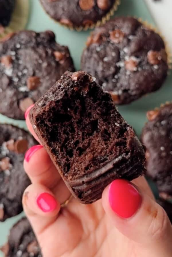 A hand with pink nail polish holds a partially eaten chocolate muffin, with more chocolate muffins visible in the background.