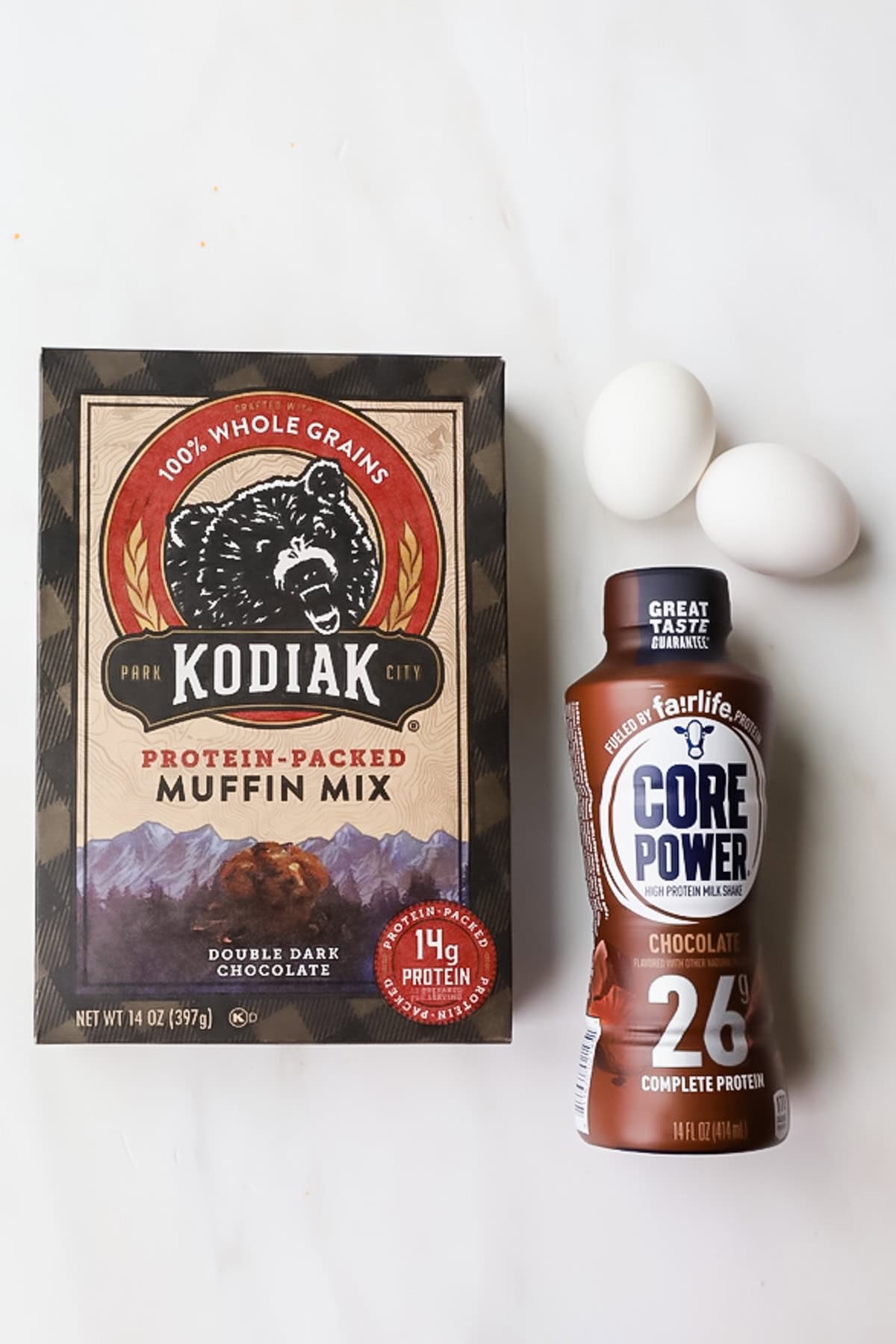 A box of Kodiak protein-packed muffin mix, a bottle of Core Power chocolate protein shake, and three white eggs are arranged on a white surface.
