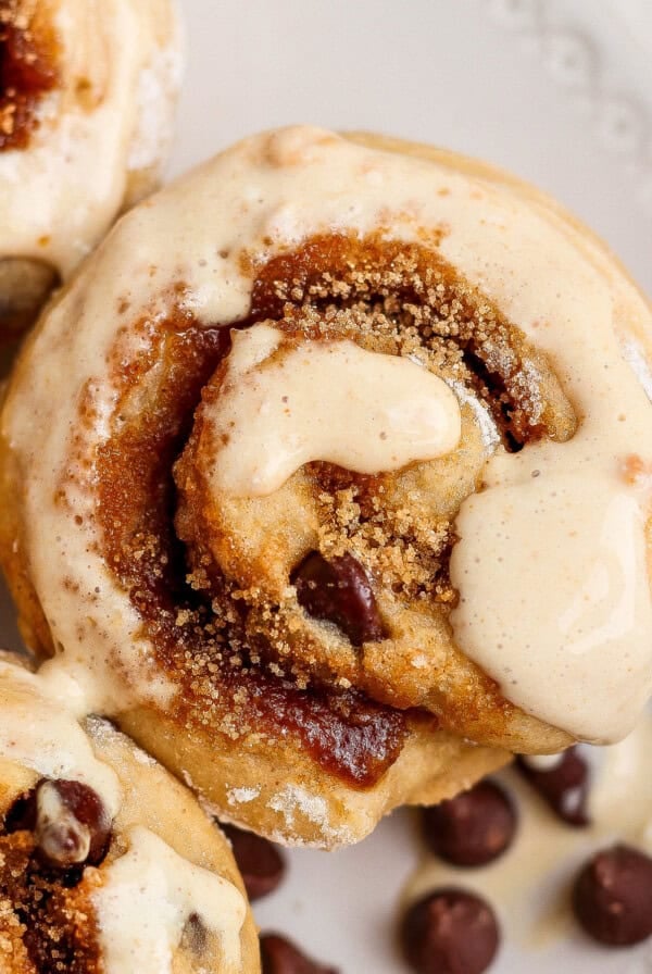 Close-up of cinnamon rolls topped with icing and sprinkled with brown sugar, with chocolate chips scattered on a white plate.