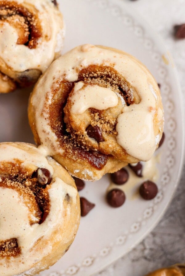 Close-up of cinnamon rolls topped with icing on a decorative white plate, with several chocolate chips scattered nearby.