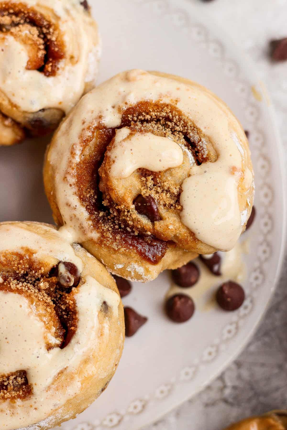 Close-up of cinnamon rolls topped with icing on a decorative white plate, with several chocolate chips scattered nearby.