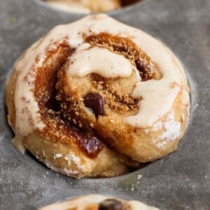 Close-up of cinnamon rolls with icing in a metal baking tray. The rolls are partially baked and display a swirl of cinnamon and sugar.